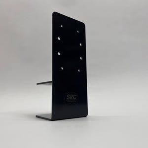 May include: A black metal bracket with a right-angle design. The bracket has a vertical plate with multiple holes and a horizontal shelf. The letters "SRC" are visible on the bracket.