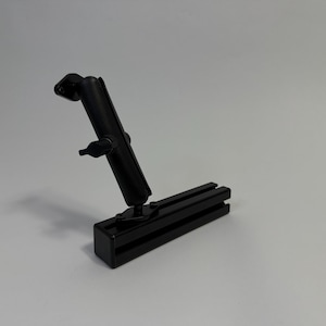 May include: A black, adjustable mounting bracket with a rectangular base and a pivoting arm. The arm has a ball joint for flexible positioning. The bracket is designed to attach to a surface and hold an object.
