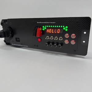 May include: A black racing simulator control panel with multiple buttons, switches, and a digital display showing "88888". The panel has the text "SIMRACINGCRAFTSMEN" printed on it. Features red and green lights.