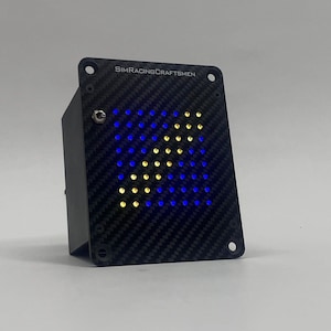May include: A black rectangular device with a carbon fibre texture on the front panel. The panel displays a blue and yellow arrow made of LED lights. The text "SIMRACINGCRAFTSMEN" is printed at the top.