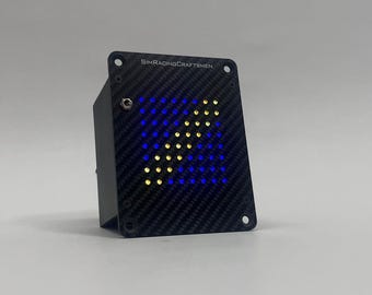 May include: A black rectangular device with a carbon fiber texture on the front panel. The panel displays a blue and yellow arrow made of LED lights. The text "SIMRACINGCRAFTSMEN" is printed at the top.