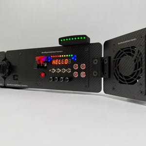 以下が含まれることがあります： カーボンファイバーの質感を持つ黒い電子デバイス。ボタン、ライト、デジタルディスプレイを備えています。ファンと一連の緑色のライトが含まれています。「SR Racing Craftsmen」の文字が見えます。