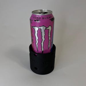 May include: A pink and silver Monster Energy drink can sits inside a black can cooler. The can has the words "ZERO SUGAR" printed on it. The cooler has a cylindrical shape with several small holes around the sides.