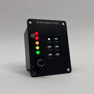 May include: A black carbon fiber sim racing control panel with the text "SIMRACINGCRAFTSMEN". It features six buttons labeled "SRC", a rotary dial, and a series of colored LED lights: red, orange, yellow, and green.