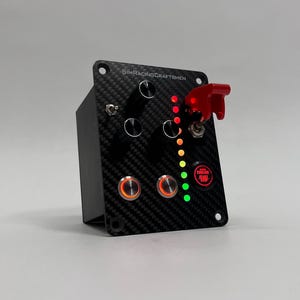 May include: A black control panel with a carbon fiber texture, featuring buttons, switches, and indicator lights. The panel has a red toggle switch and the text "SIMRACINGCRAFTSMEN". The panel also has a red button with the text "FUELING PUMP".