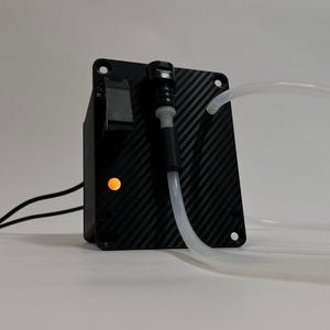 以下が含まれることがあります： カーボンファイバー柄の黒い長方形のコントロールボックスで、四隅に取付穴があります。黒いトグルスイッチ、点灯したオレンジ色のインジケーターライト、黒いポートに接続された透明なプラスチックチューブが特徴です。