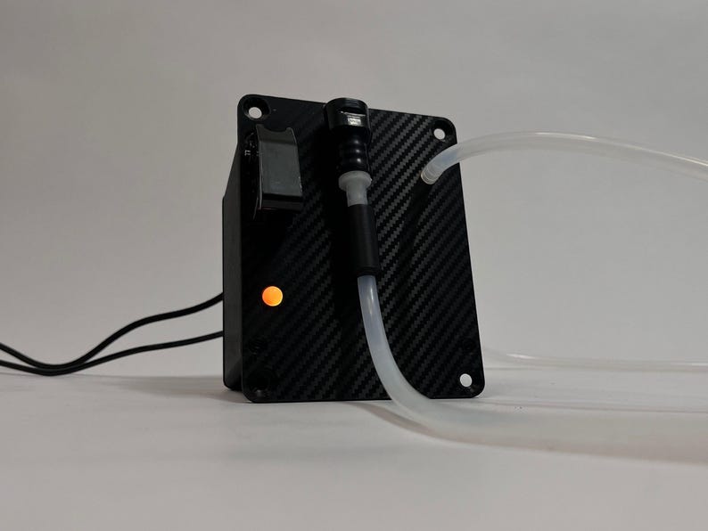 May include: A black carbon fiber patterned control box featuring a rectangular shape with four corner mounting holes. It includes a black toggle switch, a glowing orange indicator light, and a clear plastic tube connected to a black port.
