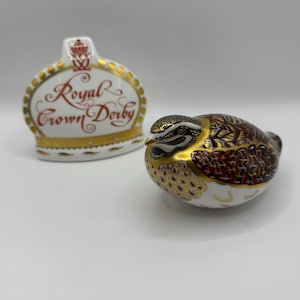 May include: A Royal Crown Derby trinket box with a crown-shaped lid, featuring the words "Royal Crown Derby" in red script. A decorative bird figurine with brown, gold, and white detailing sits beside it.