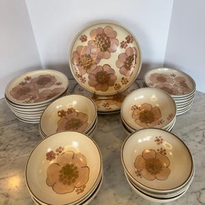 Puede incluir: Un juego de vajilla de cerámica vintage con estampado floral. El juego incluye platos, cuencos y una fuente para servir. El estampado floral es en tonos de rosa, marrón y verde sobre un fondo crema.