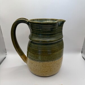 Puede incluir: Una jarra de cerámica hecha a mano con un asa grande y un pico. La jarra es principalmente verde con una base beige y una superficie texturizada. El diseño presenta una textura acanalada alrededor del centro.