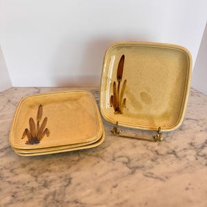 Può includere: Set di quattro piatti quadrati in ceramica con una glassa giallo chiaro e un motivo floreale marrone. I piatti sono impilati uno sopra l'altro.