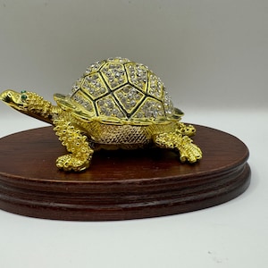 May include: A gold turtle figurine with a shell covered in rhinestones. The turtle is sitting on a wooden base.