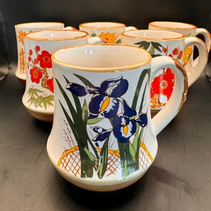 May include: Set of six white ceramic mugs with floral designs. The mugs feature different floral patterns, including red flowers, yellow flowers, and blue irises. The mugs have a brown rim and a brown base.