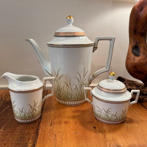 May include: A white porcelain tea set with a gold trim and a green and gold floral design. The set includes a teapot, creamer, and sugar bowl.