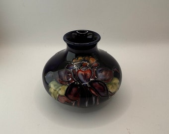 Puede incluir: Un pequeño jarrón de cerámica azul oscuro con un diseño floral. El jarrón tiene un cuerpo redondo y un cuello estrecho. El diseño floral presenta un grupo de flores en tonos rojos, morados y amarillos.