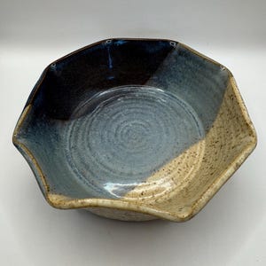 Puede incluir: Un cuenco de cerámica hecho a mano con una forma única de múltiples lados. El cuenco presenta un degradado de color, que va del azul oscuro al azul claro y al beige. El interior tiene un patrón en espiral.