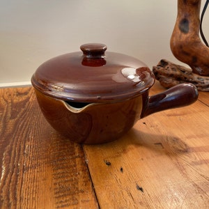 Puede incluir: Una olla de cerámica marrón con tapa y asa. La olla tiene un pico vertedor en el lateral.