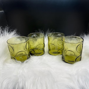 Könnte beinhalten: Vier olivgrüne Trinkgläser mit einem strukturierten, kreisförmigen Design. Die Gläser sind aus Glas und auf einer weißen, flauschigen Oberfläche angeordnet. Die Gläser sind leer und bereit zum Befüllen.