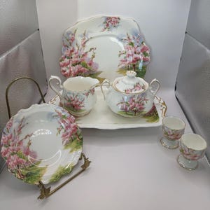 Könnte beinhalten: Vintage Porzellan-Teeservice mit rosa Blütenmuster. Das Set beinhaltet eine Servierplatte, einen Sahnekännchen, eine Zuckerdose, zwei Eierbecher und eine Servierschüssel. Die Teile sind weiß mit goldenen Verzierungen.