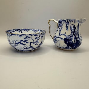 Könnte beinhalten: Ein blau-weißes Porzellan-Schalen- und Krug-Set. Die Schale hat einen gewellten Rand und ein blaues Landschaftsdesign. Der Krug hat einen Griff mit goldenem Besatz und eine blaue Illustration einer Person. Beide Teile haben einen goldenen Besatz.