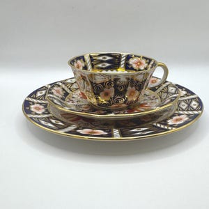 Könnte beinhalten: Ein dreiteiliges Teeservice mit einer Teetasse, Untertasse und Teller. Das Set hat einen marineblauen Hintergrund mit goldenen Verzierungen und floralen und geometrischen Mustern in Orange und Weiß. Die Teetasse hat einen goldenen Henkel.