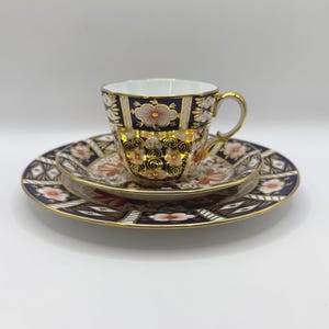 May include: A three-piece tea set featuring a teacup and two plates. The set has a navy blue background with gold trim and floral patterns in shades of pink and orange. The teacup has a gold handle.
