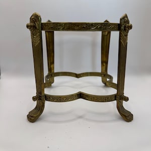 Może przedstawiać: Zabytkowy, mosiężny, dekoracyjny stojak z misternymi detalami. Stojak posiada kwadratowy blat, cztery pionowe podpory i ozdobne, zakrzywione podpory podstawy. Mosiądz ma postarzaną patynę. Stojak przeznaczony jest do przechowywania małego przedmiotu.