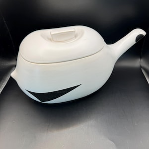Pode incluir: Bule de cerâmica branco com tampa e alça. O bule tem um design de triângulo preto na lateral e um ponto preto na alça. O bule está em uma superfície escura.
