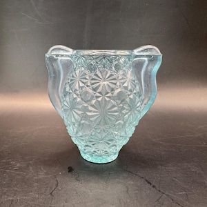 Op de afbeelding: Een lichtblauwe glazen vaas in vintage-stijl met een gestructureerd sterrenpatroon. De vaas heeft een uitlopende vorm met twee gebogen handvatten aan weerszijden. Het glas heeft een lichte iriserende glans.