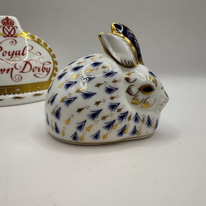 Puede incluir: Figura de conejo de cerámica blanca con detalles azules y dorados. El conejo presenta un patrón de formas de hojas azules y doradas. Una segunda pieza de cerámica con las palabras "Royal Crown Derby" está en el fondo.