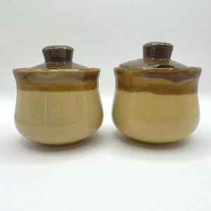 May include: Two ceramic jars with lids. The jars are a light tan colour with a darker brown band around the top and a brown lid. The jars are round with a slight curve and a small handle on the lid.