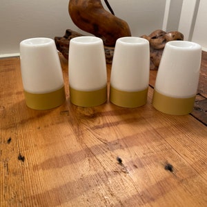 May include: Four white ceramic egg cups with a gold band around the bottom. The cups are arranged in a row on a wooden surface.