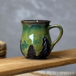 Peut inclure: Une tasse en céramique faite à la main avec un motif de forêt. La tasse présente un glaçage vert avec des silhouettes d'arbres brun foncé. La poignée est incurvée et la tasse repose sur une surface en bois. La tasse mesure environ 10 cm de haut.