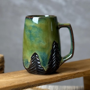 May include: A green ceramic mug with a forest design. The mug has a dark brown handle and a textured black tree design at the base. The mug is sitting on a wooden tray.