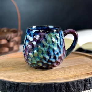 Peut inclure: Une tasse en céramique faite à la main avec une surface texturée, aux teintes de bleu, de violet et de vert. La tasse a une anse foncée et repose sur un plateau de service en bois avec un bord foncé et texturé. Un panier et un livre ouvert sont en arrière-plan.