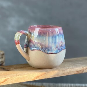 Könnte beinhalten: Eine handgefertigte Keramiktasse mit rundem Boden und Henkel. Die Tasse hat eine cremefarbene untere Hälfte und eine obere Hälfte mit einer Mischung aus rosa, blau und lila Glasur. Die Tasse steht auf einer Holzoberfläche.