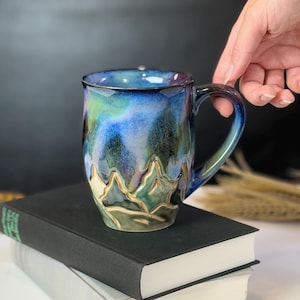 May include: A handmade ceramic mug with a blue, green, and purple glaze. The mug features a mountain-like design around the base and is held by a hand. The mug is sitting on top of two books.