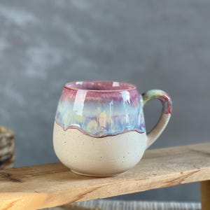 Könnte beinhalten: Handgefertigte Keramiktasse mit abgerundeter Form und gesprenkelter, cremefarbener Basis. Der obere Teil der Tasse hat eine Glasur mit rosa, blauen und grünen Farbtönen. Der Henkel ist hellgrün.