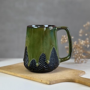 Könnte beinhalten: Eine grüne Keramiktasse mit einem Walddesign. Die Tasse hat eine dunkelgrüne Glasur mit einem schwarzen Baumdesign, das in die untere Hälfte geschnitzt ist. Die Tasse steht auf einem Holzschneidebrett.