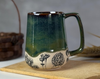 May include: A handmade ceramic mug with a dark green glaze and a brown rim. The lower portion of the mug features black and white illustrations of various mushrooms. The mug is set on a wooden chopping board.