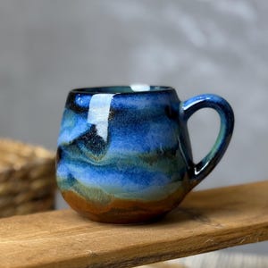Könnte beinhalten: Handgefertigte Keramik-Tasse mit rundem Korpus und kleinem Henkel. Die Glasur weist Schattierungen von Blau, Grün und Braun auf, wodurch ein geschichteter, ozeanähnlicher Effekt entsteht. Die Tasse steht auf einer Holzoberfläche.