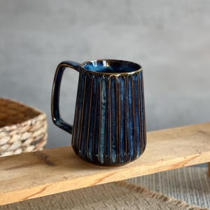 May include: A blue ceramic mug with vertical ribbed detailing. The mug has a dark blue exterior with a gold rim and handle. The mug is on a wooden surface. A woven basket and a beige placemat are in the background.