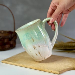 May include: A handmade ceramic pitcher with a light green and white glaze. The pitcher has a curved handle and is held over a wooden cutting board. The glaze has a watercolor effect, with streaks of color.