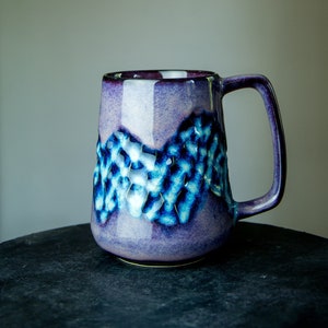 Könnte beinhalten: Eine Keramiktasse mit strukturiertem Design in Lila- und Blautönen. Die Tasse hat einen Henkel und eine weite Öffnung. Das Design weist ein strukturiertes Muster in Blau- und Weißtönen mit lila Basis und Rand auf.