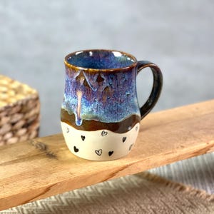 May include: A ceramic mug featuring a two-tone design with a glossy, mottled blue and purple glaze on the upper portion and a cream-colored base decorated with small, hand-painted black heart outlines. The mug has a curved handle and a smooth finish.