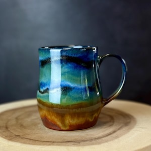 May include: A handmade ceramic mug with a unique, colorful glaze. The mug features a blend of blues, greens, and browns, creating a marbled effect. The mug has a curved handle and is set on a wooden surface. Perfect for coffee or tea.