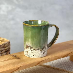Könnte beinhalten: Ein Keramikbecher mit einer glänzenden grünen Glasur im oberen Bereich, die in eine gesprenkelte weiße Basis übergeht. Der Becher hat einen geschwungenen Henkel auf der rechten Seite und einen glatten, zylindrischen Körper mit einem Tropfmuster.