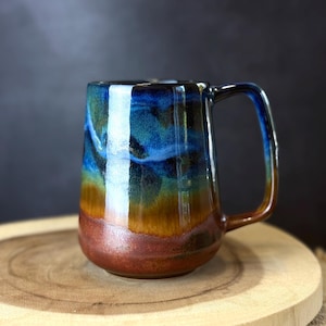 Op de afbeelding: Een handgemaakte keramische mok met een uniek glazuur. De mok heeft een kleurengradiënt, waaronder blauw, bruin en groen. De mok heeft een grote handgreep en een afgeronde basis. De mok staat op een houten oppervlak.