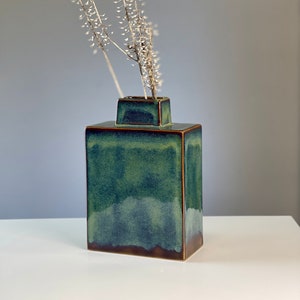 Pode incluir: Um vaso quadrado de cerâmica com um esmalte verde escuro e uma borda marrom. O vaso está cheio de flores brancas secas.