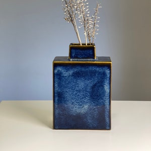 Könnte beinhalten: Eine dunkelblaue Keramikvase mit goldfarbenem Rand und quadratischer Form. Die Vase ist mit getrockneten weißen Blumen gefüllt.